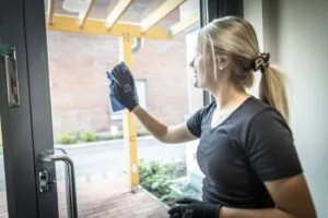 Person med hansker tørker glassdør med blå klut innendørs.