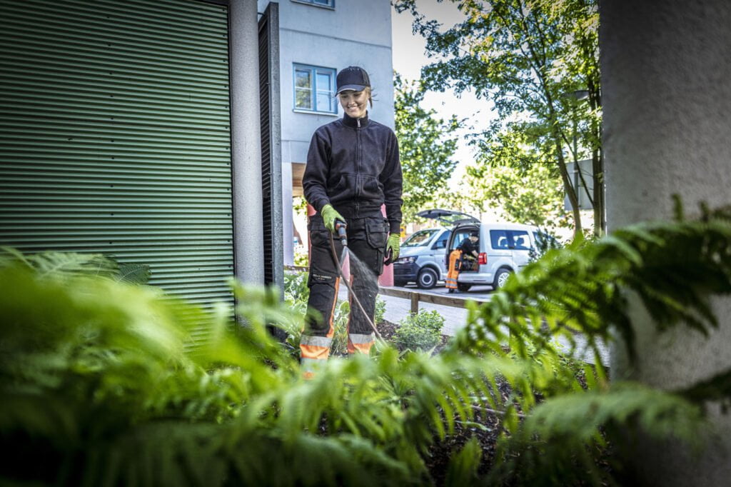Person med hansker sprøyter planteområde foran bygning, bil i bakgrunnen.