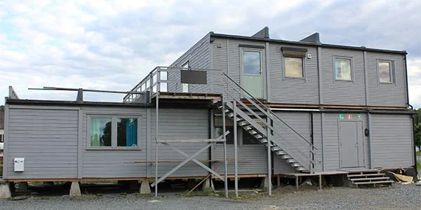 Grå modulbygg med utvendig trapp og metallrekkverk under skyet himmel.
